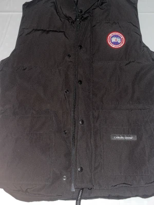 Svart dunväst från Canada Goose - Svart dunväst från Canada Goose med klassisk logga på bröstet och två stora fickor framtill. Västen har dragkedja och knappar samt diskret Canada Goose-märke på ena fickan. Perfekt för kalla dagar när du vill hålla värmen och ändå se stilren ut.
