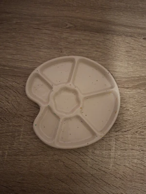 Vit målarpalett i plast - En vit målarpalett i plast med sex fack runt kanten och ett större fack i mitten. Paletten har en klassisk oval form med tumgrepp och är lätt att använda för att blanda färger vid målning.