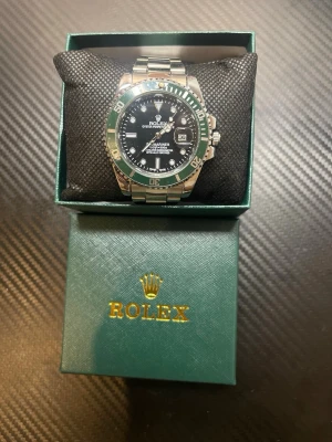 Rolx Submariner klocka grön/silver - Rolx Submariner herrklocka med grön vridring och svart urtavla. Boett och länk i rostfritt stål, datumfönster och tydliga indexmarkeringar. Kommer i  grön Rolx-box med guldfärgad logga.