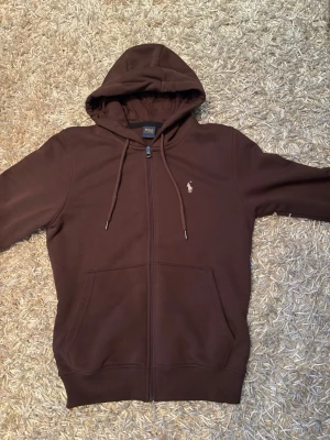 Brun Ralph lauren kofta  - Mörkbrun hoodie från Polo Ralph Lauren med dragkedja, huva med dragsko och två stora fickor framtill. Klassisk broderad logga på bröstet. Tillverkad i mjukt bomullsmaterial som känns skönt mot huden.