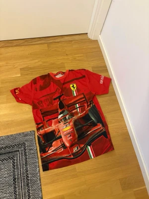 Röd Ferrari F1 t-shirt - Snygg röd t-shirt med stort Ferrari Formel 1-motiv på framsidan och klassiska Ferrari-loggan på ryggen. T-shirten har korta ärmar och tryckta detaljer, inklusive racingbil och text på ärmen. Perfekt för motorsportfans som vill sticka ut.