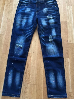 Mörkblå distressed jeans med färgstänk - Säljer ett par mörkblå jeans från MSU2 med slitna detaljer och färgstänk för en cool och trendig look. Jeansen har raka ben, klassisk femficksmodell och kontrastsömmar. Perfekta för dig som gillar streetstyle och vill sticka ut lite extra.