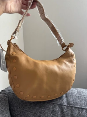 Beige axelremsväska från Daniel Ray - Snygg beige handväska från Daniel Ray i mjukt material med rundad form. Väskan har dekorativa runda nitar längs kanterna och en bred axelrem. Perfekt för dig som vill ha en trendig accessoar med plats för det viktigaste. 30x20x6 cm 