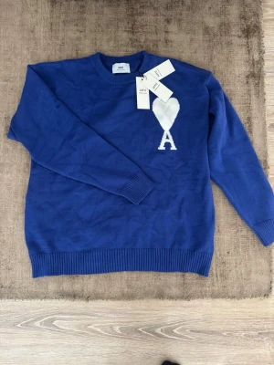 Ami tröja  - Snygg blå stickad tröja från Ami med rund halsringning och ribbade muddar. På bröstet finns ett vitt hjärta och bokstaven A som ger en unik touch. Perfekt för dig som vill ha något stilrent men ändå sticker ut. Fint skick, sitter som en stor S eller m. Lite större i storlek. Tags medföljer. 