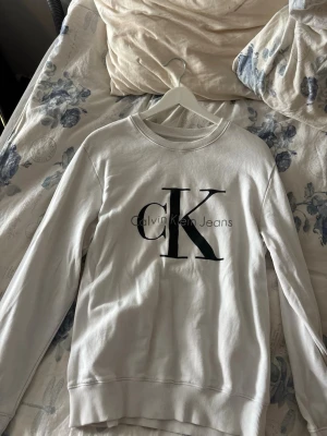 Vit Calvin Klein Jeans sweatshirt - Vit sweatshirt från Calvin Klein Jeans med stor svart CK-logga på bröstet. Tröjan har rund halsringning, långa ärmar och ribbade muddar vid ärmslut och nederkant. Perfekt för en clean och stilren look.