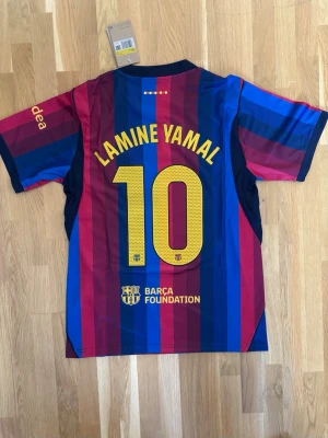 Barcelona matchtröja Lamine Yamal #10 - Säljer en officiell FC Barcelona matchtröja med Lamine Yamal och nummer 10 på ryggen. Tröjan har klassiska blå och röda ränder, Nike-logga och Barca-emblem på bröstet samt tryck i gult. Perfekt för dig som älskar fotboll och vill stötta ditt lag.