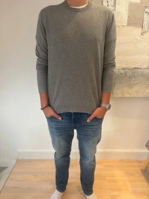 Grå stickad tröja med rund hals - En enkel och stilren grå stickad tröja med rund halsringning och långa ärmar. Tröjan har en rak passform och är tillverkad i ett mjukt, tunt material som passar perfekt till jeans. Perfekt för dig som gillar minimalistisk stil.