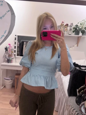 Blåvitrutig topp med volang - Supersöt topp i blåvitt rutigt mönster med puffärmar och volang nertill. Modellen är kortare och har en feminin, figurnära passform. Perfekt för dig som gillar en söt och trendig stil. Materialet känns lätt och luftigt.