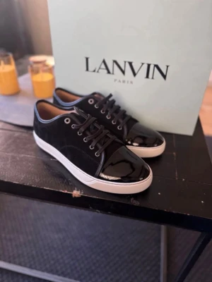 Svarta sneakers från Lanvin - Snygga svarta sneakers från Lanvin med blank tå i lack och ovandel i mocka. Skorna har vita sulor, svarta skosnören och metallfärgade öljetter. Perfekta för dig som vill ha en clean och exklusiv look.