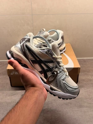 Asics- Gel kayano 14 - Snygga sneakers från Asics i vitt och silver med svarta detaljer. Ovandelen är i mesh och syntetmaterial för en sportig vibe. Skorna har en chunky sula och klassisk Asics-logga på sidan. Perfekta för dig som gillar streetwear och sportig stil.