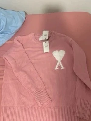 Rosa stickad Ami tröja  - Supersöt rosa stickad tröja med vit hjärtdetalj och bokstaven A på bröstet. Tröjan har rund halsringning, långa ärmar och breda ribbade muddar vid ärmslut och nederkant. Perfekt för dig som vill ha en mysig och trendig look.