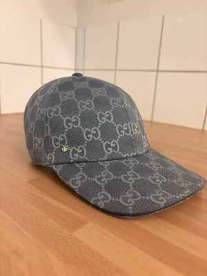 Blå keps med GG-mönster från Gucci - Snygg blå keps från Gucci med det klassiska GG-monogrammet över hela kepsen. Tillverkad i ett slitstarkt tyg med justerbart spänne bak för perfekt passform. Perfekt accessoar för dig som vill sticka ut och visa stil.