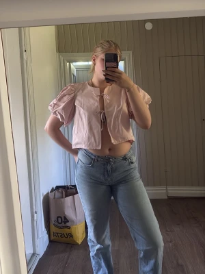 Ljusrosa croppad blus med puffärm - Söt ljusrosa blus med korta puffärmar och croppad passform. Toppen har knytning framtill och är tillverkad i ett lätt bomullstyg. Perfekt för en trendig och avslappnad look.