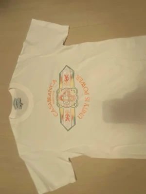 Vit Casablanca t-shirt med tryck - Säljer en vit t-shirt från Casablanca med färgglatt tryck på bröstet där det står 'CASABLANCA TENNIS CLUB' och 'UNITY IS POWER'. T-shirten har korta ärmar och en klassisk passform. Perfekt för dig som gillar stilrena men ändå unika plagg.