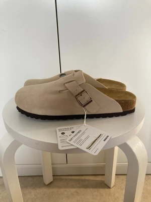 Beige Birkenstock tofflor mocka - Säljer ett par snygga beige tofflor från Birkenstock i mjuk mocka med justerbart spänne i metall. De har en klassisk slip-in design och bekväm fotbädd i kork. Perfekta för chill dagar hemma eller när du vill ha det extra bekvämt.