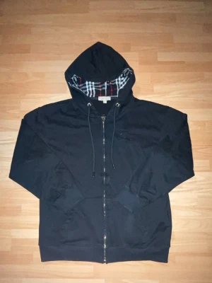 Svart burberry hoodie med dragkedja  - Svart burberry hoddie dragkedja och huva med rutigt foder i rött, vitt och grått. Justerbara snören i huvan och fickor framtill. Perfekt för en avslappnad och stilren look.