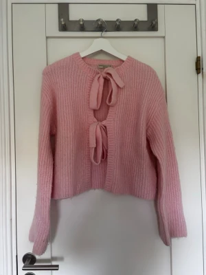 Rosa stickad kofta med knytband - Söt rosa stickad kofta med två knytband framtill. Koftan har lång ärm, ribbstickad struktur och en croppad passform. Perfekt för att lägga till en gullig touch till din outfit.