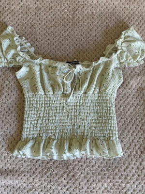 Ljusgrön blommig blus med volang - Supersöt ljusgrön blus från Gina  med små blommor, puffärmar och volangkant. Smockad midja för en tight passform och knut framtill. Aldrig använd. Perfekt för sommaren!