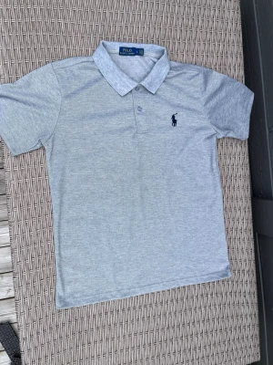 Ljusgrå pikétröja från Polo Ralph Lauren - Snygg ljusgrå pikétröja från Polo Ralph Lauren med klassisk krage och korta ärmar. Tröjan har två knappar vid halsen och den ikoniska mörkblå loggan broderad på bröstet. Perfekt till jeans eller shorts för en clean och stilren look.