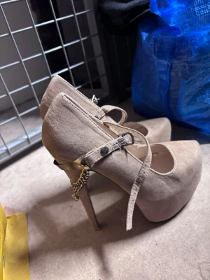 Beige platåpumps med kedjedetalj - Säljer ett par beigea pumps med hög platå och smal klack. Skorna har en Mary Jane-rem med knapp och en snygg guldfärgad kedjedetalj vid hälen. Materialet ser ut att vara mockaimitation och de har rundad tå. Perfekta för dig som vill sticka ut.
