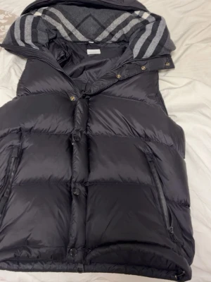 Svart burberry väst/jacka  - Snygg svart dunväst med vaddering och avtagbar huva. Insidan av huvan har ett grått och vitt rutigt mönster. Västen har dragkedja och knappar framtill samt två fickor med dragkedja. Perfekt för lager på lager och håller dig varm under kyliga dagar.