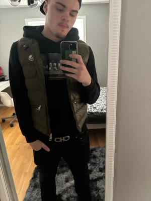Olivgrön dunväst från Moncler - Snygg olivgrön dunväst från Moncler med klassisk logga på bröstet och dragkedja framtill. Västen har en quiltad design och är tillverkad i glansigt material som ger en exklusiv känsla. Perfekt att bära över en hoodie för en trendig streetwear-look.