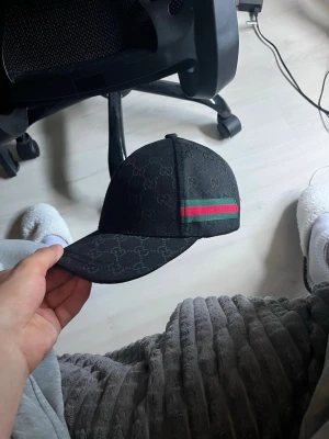 Svart Gucci keps med GG-mönster - Snygg svart keps från Gucci med det klassiska GG-mönstret över hela kepsen. På sidan finns en grön och röd rand som ger en ikonisk touch. Kepsen har böjd skärm och justerbar rem bak. Perfekt för dig som vill ha en stilren accessoar.