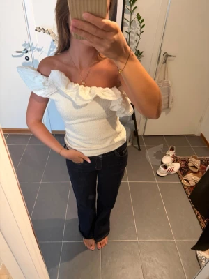 Vit offshoulder topp med volang - Säljer en superfin vit offshoulder topp med stor volangdetalj längs urringningen. Toppen är ärmlös och har en tight passform som framhäver figuren. Perfekt för dig som vill ha något trendigt och unikt i garderoben!