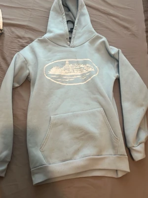 Ljus blå tröja från corteiz - Ljusblå hoodie från corteiz med stor ficka framtill och huva. Hoodien har ribbade muddar vid ärmar och nederkant. Perfekt för en avslappnad och chill stil.
