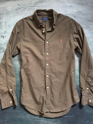Olivgrön skjorta från Polo Ralph Lauren - Säljer en olivgrön långärmad skjorta från Polo Ralph Lauren i slim fit-modell. Skjortan har klassisk krage, knappar framtill och den ikoniska broderade loggan i orange på bröstet. Perfekt för dig som gillar stilrena plagg med snygga detaljer.