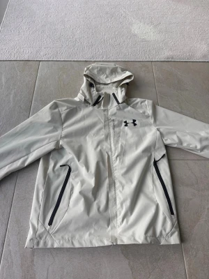Vit vindjacka från Under Armour - Snygg vit vindjacka från Under Armour med svart logga på bröstet och svarta dragkedjor på fickorna. Jackan har huva och hel dragkedja framtill. Perfekt för blåsiga dagar och enkel att matcha med olika outfits.