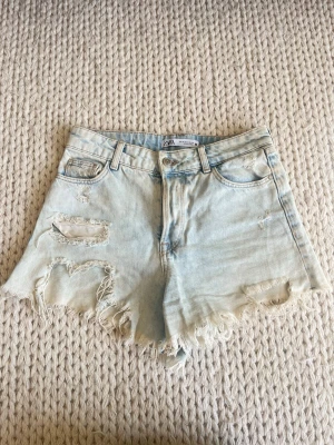 Ljusblåa denimshorts från ZARA - Säljer ett par ljusblåa denimshorts från ZARA med hög midja och råa, fransiga kanter. Shortsen har slitna detaljer framtill och klassisk femficksdesign. Perfekta för varma dagar och ger en avslappnad vibe.