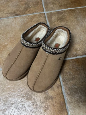 Bruna  low  UGG tofflor  - Mysiga bruna tofflor från UGG i mocka med fluffigt vitt fårskinnsfoder. Tofflorna har en dekorativ kant i svart, vitt och beige mönster runt öppningen. Perfekta för att hålla fötterna varma hemma.