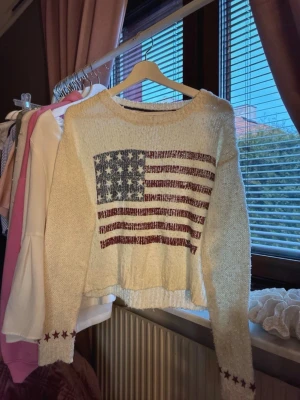 Vit stickad tröja med USA-flagga - Stickad vit tröja från KappAhl med USA-flagga framtill. Tröjan har lång ärm och röda stjärnor vid ärmsluten. Perfekt för dig som gillar en avslappnad och trendig look.🌸