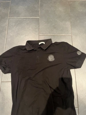Svart pikétröja från Moncler - Snygg svart pikétröja från Moncler med klassisk krage och knappar framtill. Tröjan har Moncler-logga på bröstet och ärmen. Perfekt till en stilren och avslappnad look. Passar L/M
