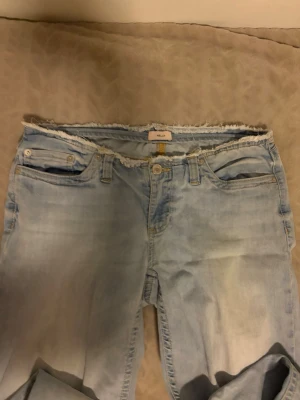 Ljusblå jeans med råa kanter - Säljer ett par ljusblå jeans från Nelly med trendiga råa kanter i midjan och vid bensluten. Jeansen har klassisk femficksdesign och raka ben. Perfekta för dig som gillar en avslappnad och cool stil.