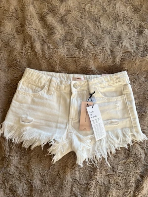 Vita lågmidjade jeansshorts - Säljer ett par vita jeansshorts med fransig kant och slitna detaljer framtill. Helt oanvända endast testade 