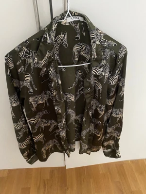 Grön zebramönstrad skjorta - Snygg grön skjorta med zebramönster och knappar framtill. Skjortan har lång ärm och är tillverkad satin , lätt material som ger en avslappnad känsla. Perfekt för dig som vill sticka ut med ett djuriskt print.