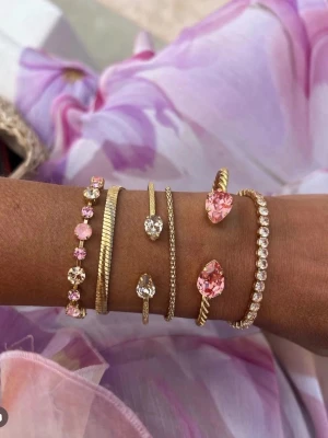 Maria svedbom armand  - Snyggt armband i guldfärgad metall med två droppformade rosa stenar i varje ände. Armbandet är öppet och har en tunn, elegant design som passar perfekt till en trendig stil. Stenarna är facetterade och ger ett lyxigt intryck.