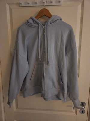 Olivia Rodrigo Ljusblå hoodie - Säljer en ljusblå hoodie med dragkedja framtill och stor huva. På ryggen finns ett stort svart tryck av ett konstnärligt ansikte. Hoodien har fickor på sidorna och ribbade muddar vid ärmar och nederkant. Perfekt för en chill och avslappnad stil.