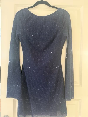 Mörkblå glittrig klänning med öppen rygg - Supersnygg mörkblå kortklänning med glitterdetaljer och lång ärm. Klänningen har en djup öppen rygg och lätt utsvängda ärmar. Använts 2 gånger! 🥰