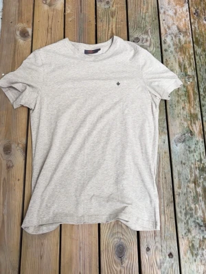 Beige Morris T-shirt  - Säljer en stilren beige t-shirt i mjuk bomull. T-shirten har rund halsringning, korta ärmar och en liten diskret broderad detalj på bröstet. Perfekt basplagg för alla tillfällen och enkel att matcha med andra plagg.