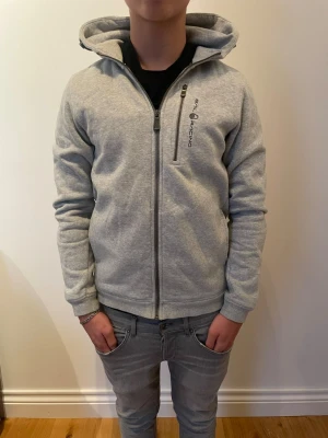 Sail racing zip hoodie - Ljusgrå hoodie med dragkedja från Sail Racing. Tröjan har huva, ribbade muddar och logga tryckt på bröstet samt en liten logga på huvan. Tillverkad i mjukt sweatshirtmaterial, perfekt för en avslappnad stil. Tröjan sitter väldigt bra om man e runt 176-180