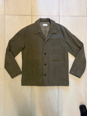 Overshirt These Glory Days herr  - Stilren olivgrön overshirt i kraftigt bomullstyg med tre stora utanpåliggande fickor framtill och knäppning med fyra knappar. Klassisk krage och raka ärmar med knappdetaljer vid ärmslut. Perfekt lager på lager-plagg för en avslappnad look. Märket är These Glory Days. Skriv gärna vid fler funderingar:)