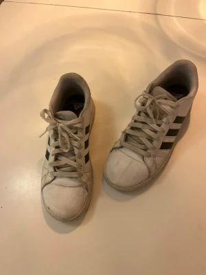 Vita Adidas sneakers med svarta ränder - Klassiska vita sneakers från Adidas med de ikoniska tre svarta ränderna på sidan. Skorna har lågt skaft, vita snören och sula i gummi. Perfekta för en avslappnad och sportig stil. Materialet är syntet och textil.
