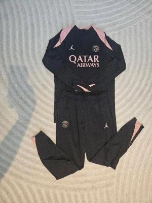 Svart PSG träningsset med rosa detaljer - Säljer ett svart PSG träningsset med långärmad tröja och byxor. Setet har rosa detaljer och loggor, samt tryck från Qatar Airways och Jordan. Perfekt för fotbollsträning eller chill. Materialet är lätt och andas bra.