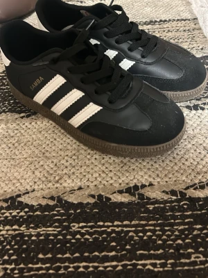 Adidas Samba svarta sneakers - Klassiska Adidas Samba sneakers i svart med vita ränder på sidorna och brun gummisula. Skorna har snörning och detaljer i både skinn och mocka för en tidlös look. Perfekta för dig som gillar streetstyle och sportig vibe.