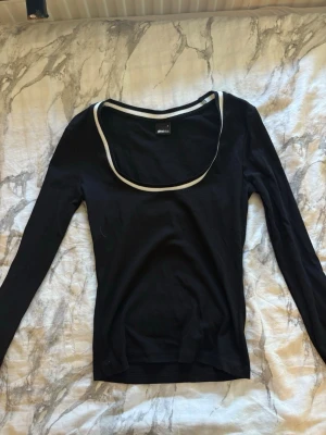 Svart långärmad topp med vit kant - Svart långärmad topp med bred, rund urringning och vit kontrastkant runt halsen. Tunn och mjuk i materialet, perfekt för lager på lager eller som enkel statement-piece. Snygg och enkel design som passar till det mesta.