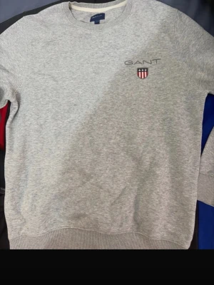 Grå sweatshirt från GANT - Klassisk grå sweatshirt från GANT med rund halsringning och broderad logotyp samt sköld på bröstet. Tröjan har ribbade muddar vid ärmslut och nederkant. Perfekt för en avslappnad och stilren look.