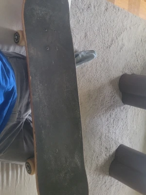 Röd skateboard med svarta griptejp - Säljer en klassisk skateboard med röd undersida och svart griptejp på ovansidan. Brädan har gulaktiga hjul och silverfärgade trucks. Formen är standard med rundad nose och tail, perfekt för tricks och cruising. Materialet är trä med sandpapprig yta på toppen.